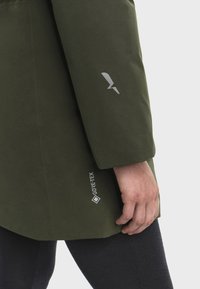 Osoba oblečená v olivovo zelenom Gore-Tex kabáte s logom na rukáve a bokoch, viditeľná ruka odpočívajúca pri boku, na jednoduchom pozadí.