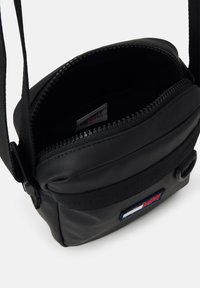Bolso de bandolera negro hecho de nailon texturizado con cierre de cremallera en la parte superior, correa ajustable y un parche con el logo en azul, blanco y rojo.