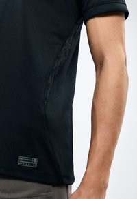 Κοντινό πλάνο ενός ατόμου που φοράει μαύρο κοντομάνικο πουκάμισο με διακριτικό πάνελ με σχέδιο και μια μικρή ετικέτα "Engineered" κοντά στο στρίφωμα.