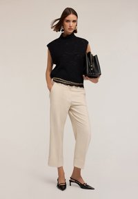 Top negru tricotat fără mâneci, cu guler înalt, pantaloni crem croiți cu manșete și sandale cu toc subțire, cu vârf ascuțit, negre. Accent din lanț de aur.