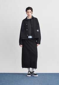 Γυναίκα που φοράει μαύρο σακάκι με κουμπιά, σκούρο γκρι πουλόβερ με πλέξη καλώδιο, λευκό πουκάμισο, μαύρη μακριά φούστα, λευκές κάλτσες και μαύρα αθλητικά παπούτσια, στέκεται σε μπλε δάπεδο.