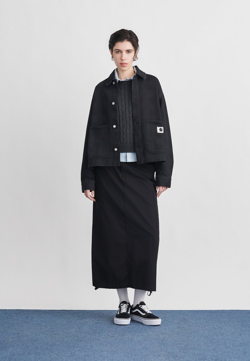 Woman wearing black button-up jacket, dark gray cable knit sweater, white shirt, long black skirt, white socks, and black sneakers standing on blue floor.