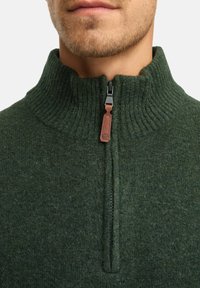 Donkergroen gebreide trui met een hoge kraag. Voorzien van een kwart ritssluiting met een bruine leren trekker en geribde structuur.