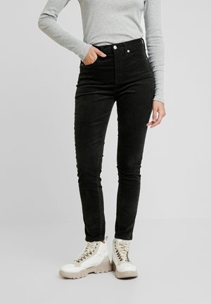 Personne portant un jean skinny noir taille haute, un haut gris à manches longues et des bottes blanches épaisses à lacets, debout avec une main dans la poche.