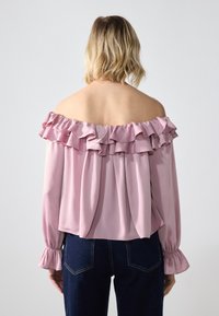 Blusa rosa scollata con colletto e polsini arricciati. Tessuto morbido e fluido con pieghe, abbinata a pantaloni blu scuro.