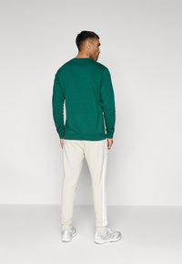 Žalia ilgam rankovėmis marškinėliai, derinami su kreminėmis kelnėmis, turinčiomis baltas juosteles šonuose, su laisvu siluetu ir minkšta tekstūra.