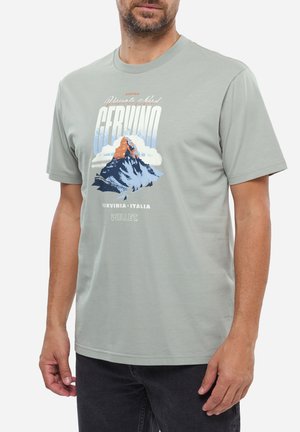 Homme portant un t-shirt gris clair avec un graphique de la montagne Cervino, des nuages et le texte "Cervinia Italia Millet" devant.
