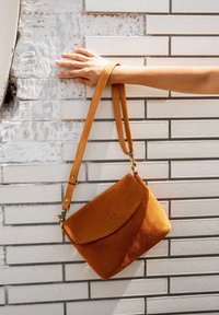 Hand mit einem hellbraunen Lederarmband, das eine braune Wildlederhandtasche gegen eine weiße geflieste Wand mit einigen abgenutzten Stellen hängen lässt.