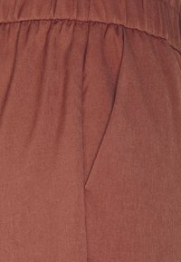 Roestkleurige stof met een gladde textuur, met geplooide details en subtiele stiksels. Inclusief een zijzak voor praktisch gebruik.