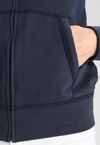 Marine blauwe zip-up hoodie met een gladde textuur, kangoeroezak en verstevigde stiksels langs de naden. Beschikt over een flexibele hemband.