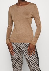 Maglione marrone a maniche lunghe con scollatura rotonda, texture a coste; abbinato a pantaloni stampati con un design geometrico nero e beige.