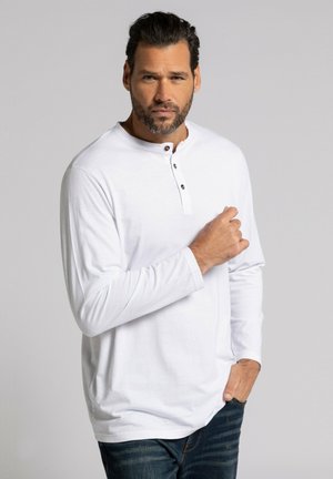 Camisa blanca de manga larga con cuello henley, que presenta tres botones oscuros y un corte relajado, combinada con unos pantalones vaqueros azules.