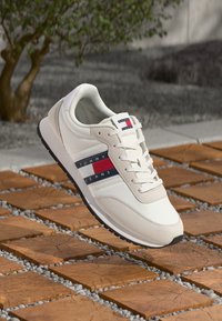 Sneaker sportivo bianco con tomaia texturizzata, chiusura con lacci e strisce blu/rosse. Suola in gomma con un accento nero, posizionata su uno sfondo di pietra.
