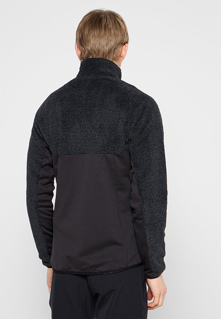 Uomo con capelli corti castano chiaro che indossa una giacca in pile nera con la parte superiore strutturata e la parte inferiore liscia, girato di spalle.