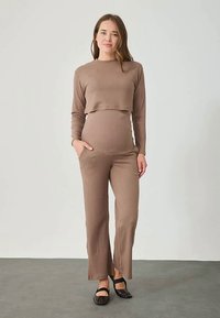 Gravid kvinna i ett ribbat, brunt matchande långärmad topp och byxor, stående med en hand i fickan och bärande svarta skor.