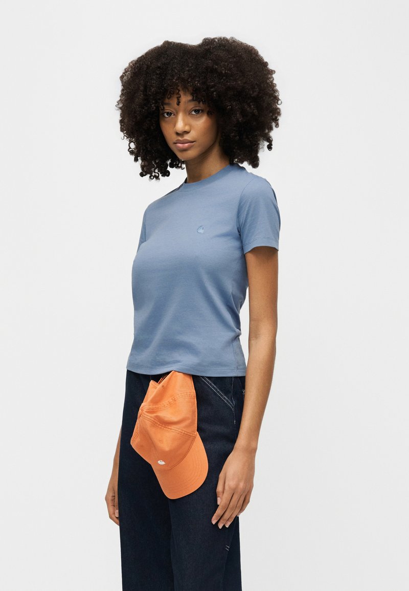 Junge Frau mit lockigem Haar trägt ein blaues, enganliegendes T-Shirt und dunkle Jeans, hält eine orangefarbene Kappe, die in ihren Hosenbund gesteckt ist.