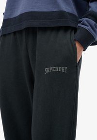 Pantalones de chándal negros hechos de una tela suave, con una cinturilla elástica, bolsillos laterales y un logo gris de "SUPERDRY" impreso en el muslo.