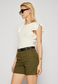 Top blanco con mangas acampanadas, combinado con pantalones cortos verde oliva. Cinturón negro con hebilla redonda. Las gafas de sol completan el conjunto.