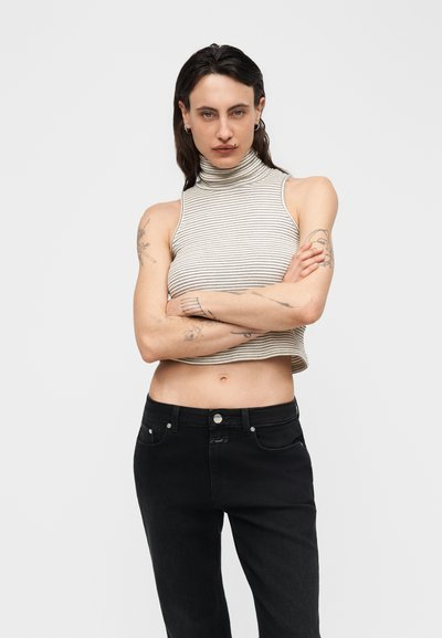 Personne aux longs cheveux foncés et tatouages, portant un crop top à col roulé rayé sans manches et un jean noir, les bras croisés, sur fond blanc.