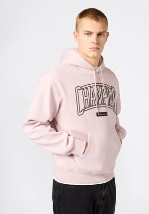 Junger Mann trägt einen hellrosa Champion-Hoodie, die Hände in der Vorderseite der Tasche verschränkt, und steht vor einem einfarbigen hellen Hintergrund.