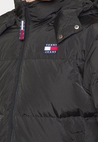 Czarna kurtka puchowa z zamkiem błyskawicznym i wysokim kołnierzem, z małą naszywką z logo Tommy Jeans na klatce piersiowej oraz guzikami na kapturze.