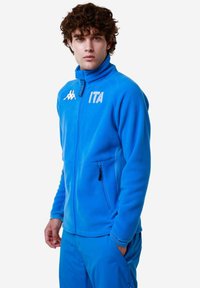 Giacca in fleece blu con collo alto, chiusura con zip e tasche laterali. Presenta un logo bianco e il testo "ITA" sul davanti. Texture liscia.