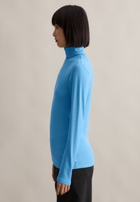 Frau mit glatten schwarzen Haaren, die einen leuchtend blauen Rollkragenpullover und schwarze Hosen trägt, steht im Profil vor einem neutralen Hintergrund.