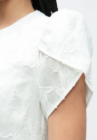 Λευκό φόρεμα με υφή, με φουσκωτά μανίκια διακοσμημένα με κεντήματα λουλουδιών και απαλή, διαφανή ύφανση, που αναδεικνύει περίτεχνα στοιχεία κατά μήκος του ώμου.