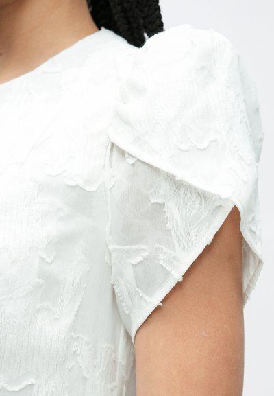 Bela obleka z teksturirano tkanino, napihnjenimi rokavi z cvetličnim vezenjem in mehkim, prosojnim materialom, ki prikazuje zapletene detajle ob rami.