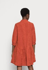 Robe couleur rouille, longueur genou, coupe décontractée, manches 3/4 boutonnées et jupe évasée. Fabriquée en tissu texturé avec un léger éclat.