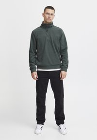Dark green fleece pullover with a zip collar, worn over a white layer, paired with black pants and white sneakers featuring black stripes.