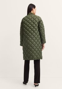 Gesteppter langer Mantel in Olivgrün mit Stehkragen, versehen mit einem Rautenmuster und langen Ärmeln, kombiniert mit schwarzen Hosen und Absätzen.