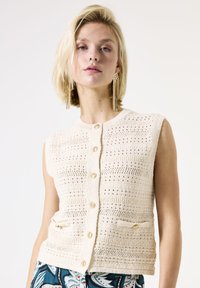 Gilet en tricot beige avec un motif texturé, fermetures à boutons dorés et deux poches avant. Design sans manches avec un col rond.