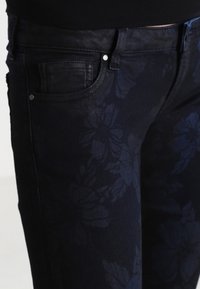 Pantalones vaqueros oscuros con un patrón floral en un tono de azul ligeramente más claro. Incluye bolsillos delanteros estándar y detalles en metal plateado.