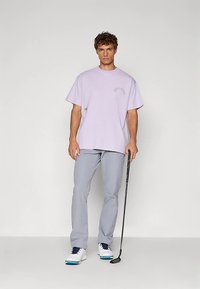 Levanduľové tričko s krátkymi rukávmi v kombinácii so svetlošedými nohavicami. Oblečenie dopĺňajú biele topánky s modrými akcentmi.