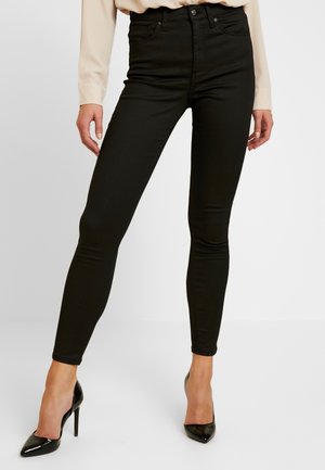 Pantalon noir ajusté porté avec un haut beige à manches longues et des chaussures à talons noirs, montré de la taille jusqu'aux pieds sur fond blanc.