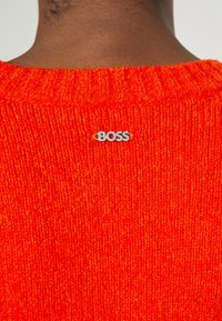 Gros plan d’un pull tricoté orange vif avec un petit logo argenté "BOSS" dans le dos près du cou.