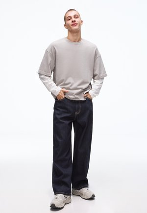 Junger Mann mit Händen in den Taschen, trägt beige und weiß geschichtete langärmlige Kleidung, dunkle weite Jeans und hellgraue Turnschuhe, stehend vor weißem Hintergrund.