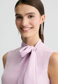 Lichtpaarse mouwloze blouse met een strikdetail bij de hals. Model draagt lange oorbellen met kristaldruppels. Glad materiaal en een zachte textuur.