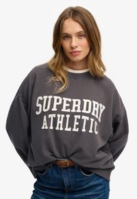 Szary sweatshirt z białym nadrukiem "SUPERDRY ATHLETIC". Charakteryzuje się luźnym krojem, okrągłym dekoltem i jest noszony na niebieskich dżinsach z plecionym paskiem.