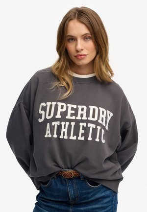 Grauer Sweatshirt mit einem weißen Druck "SUPERDRY ATHLETIC". Es hat einen lässigen Schnitt, einen Rundhalsausschnitt und wird über blauen Jeans mit einem geflochtenen Gürtel getragen.