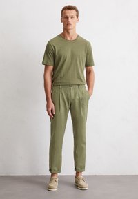 Olivengrünes T-Shirt mit Rundhalsausschnitt, kombiniert mit passenden, umgeschlagenen Hosen, beide aus leichtem Stoff. Tanfarbene Schuhe runden den Look ab.