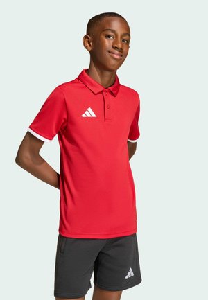 Junge steht mit den Händen hinter dem Rücken, trägt ein rotes Adidas Polo-Shirt und schwarze Adidas Shorts vor einfachem Hintergrund.