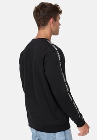 Sweatshirt noir à coupe décontractée, avec des manches longues et un imprimé logo blanc sur le côté. Tissu doux avec des poignets et un ourlet côtelés.