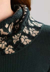 Mörkgrön ribbade tröja med en mönstrad, veckad krage i krämfärgat och mörkblå blommönster. Mjuk textur med en mysig känsla.