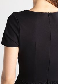 Černé šaty s krátkými rukávy, hladká tkanina, kulatý výstřih a zip na zadní straně. Obsahují strukturovaný dolní lem s geometrickým vzorem.