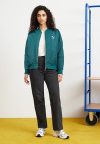 Teal bomberjakke med lynlås foran, ribbede manchetter og et lille hvidt logo. Sammen med sorte straight-leg jeans og hvide sneakers.