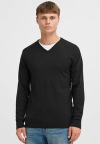 Black v-neck sweater made of a smooth knit fabric, featuring long sleeves and a subtle logo on the upper left chest. Worn over a white shirt.
