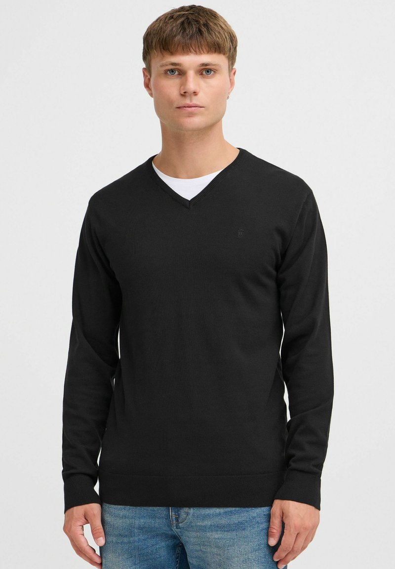 Black v-neck sweater made of a smooth knit fabric, featuring long sleeves and a subtle logo on the upper left chest. Worn over a white shirt.