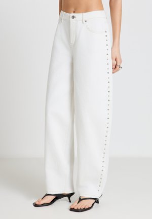 Jeans bianchi a gamba larga con borchie argento lungo le cuciture esterne, indossati con sandali neri con tacco a infradito, addome scoperto visibile su sfondo semplice.